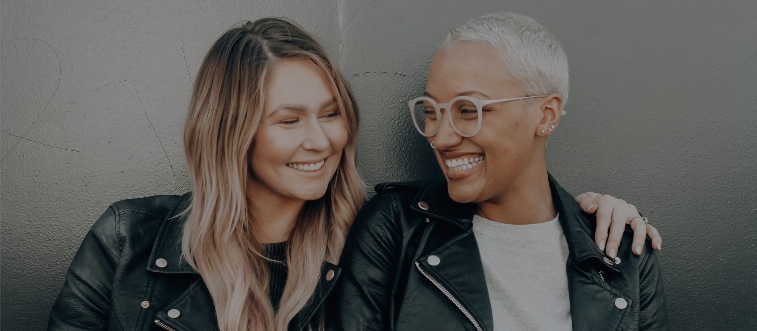 Två glada kvinnor bredvid varann lutandes mot en vägg